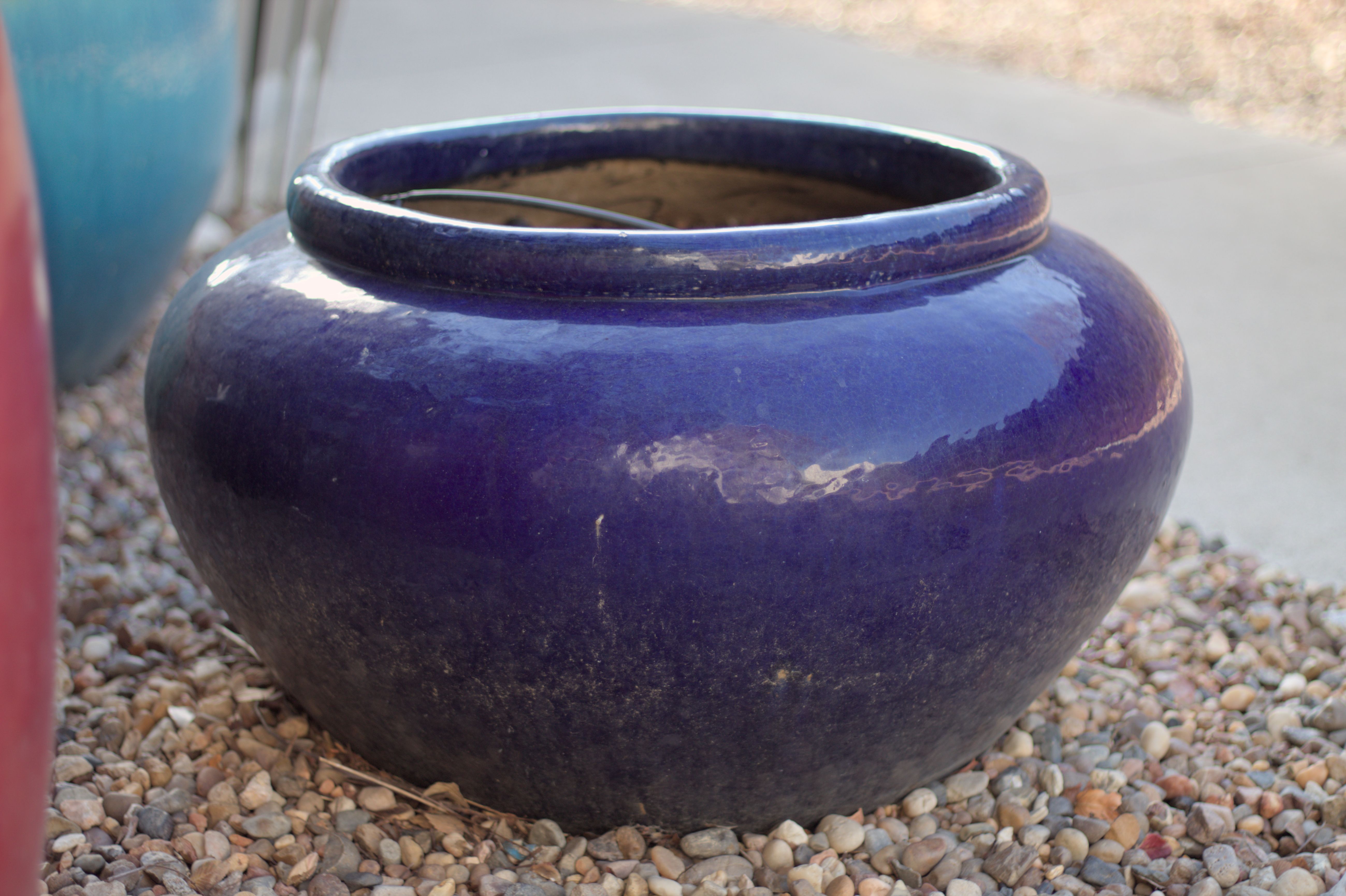 A glossy blue ceramic vase sitting on a bed of pebbles next to a concrete walkway